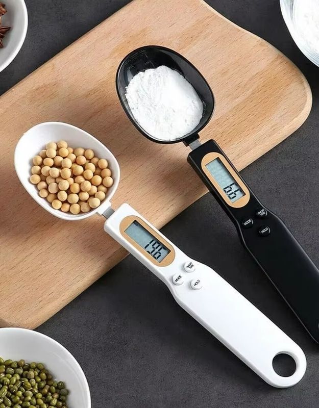A digital spoon with an LCD display showing a measurement, placed on a wooden surface with a bowl of beans beside it.